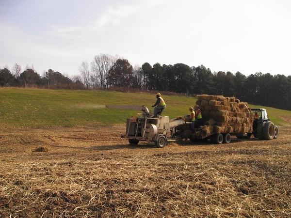 A crew applies straw to bare soil with a blower pulled by a straw supply truck.